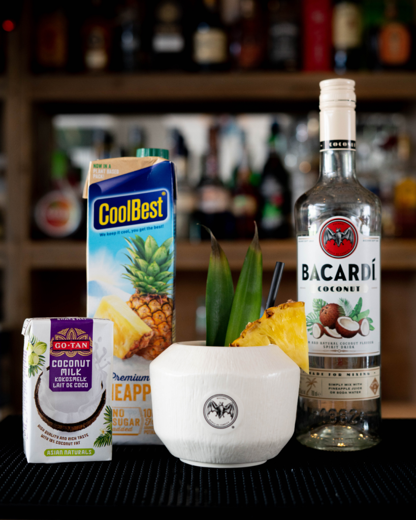 The Piña Colada on a bar top, surrounded by its ingredients.