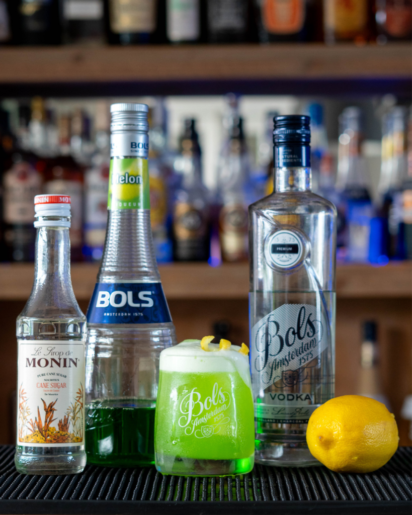 The Melon Sour on a bar top, surrounded by its ingredients.