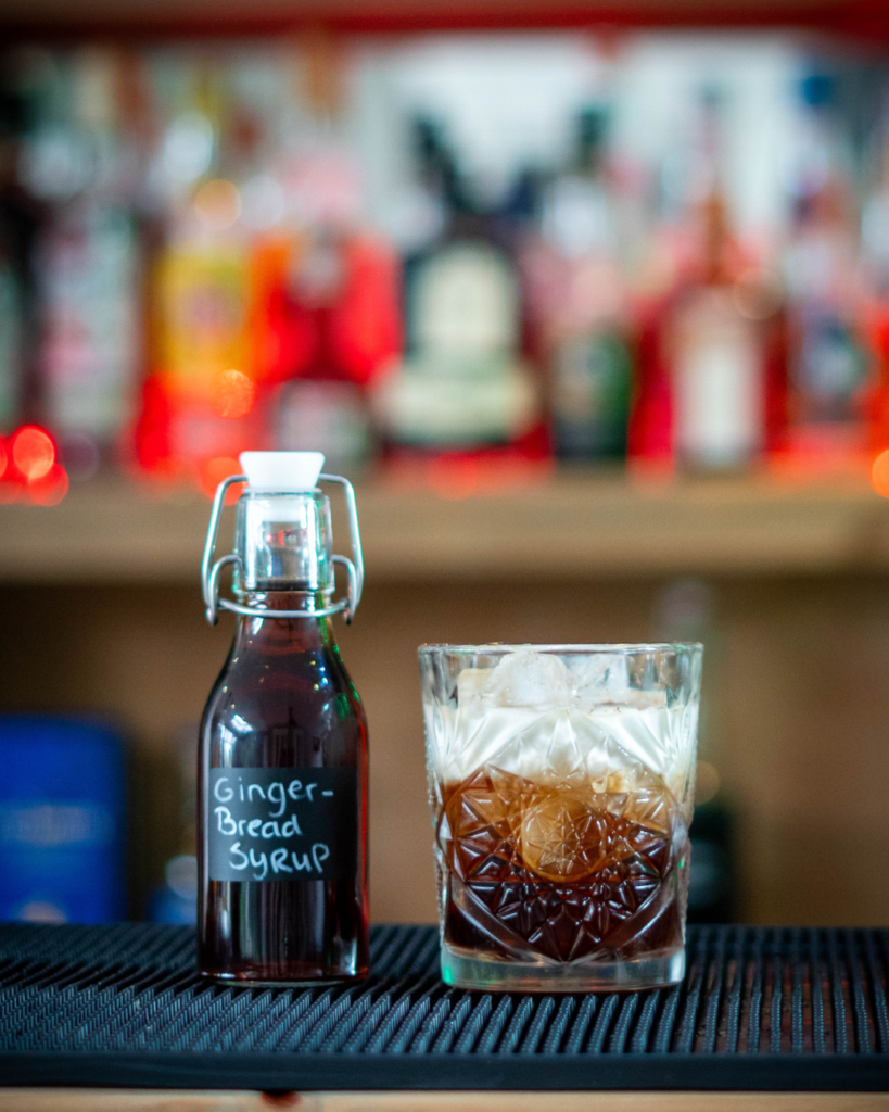 The Gingerbread White Russian and a small bottle of gingerbread syrup on a bartop, surrounded by its ingredients