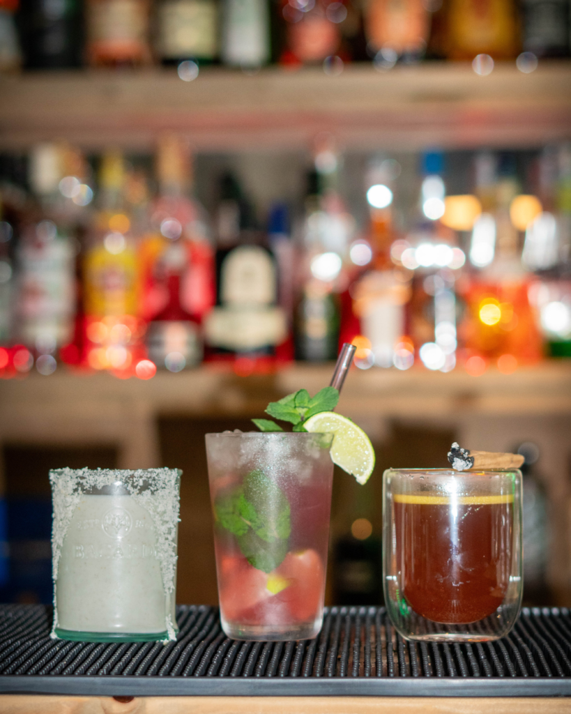 3 Rum cocktails on a bartop. From Left to Right: The Coquito, The Cranberry Mojito and The Hot Buttered Rum.