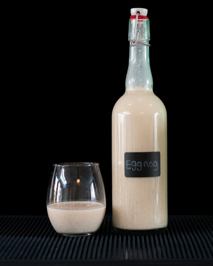 A bottle and a glass of eggnog on a bar top with black background