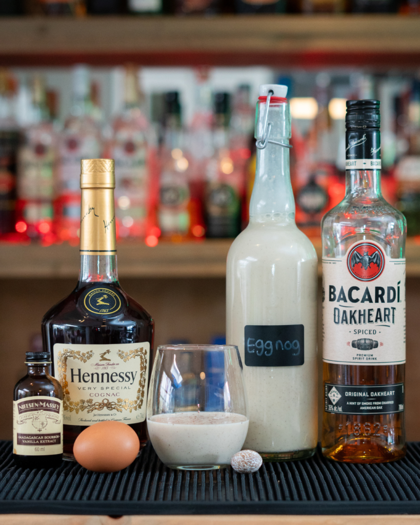 A bottle and a glass of eggnog on a bar top, surrounded by its ingredients