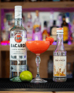 A Frozen Strawberry Daiquiri surrounded by its ingredients on a home bartop. The drink is a bright red and garnished with a fresh strawberry.