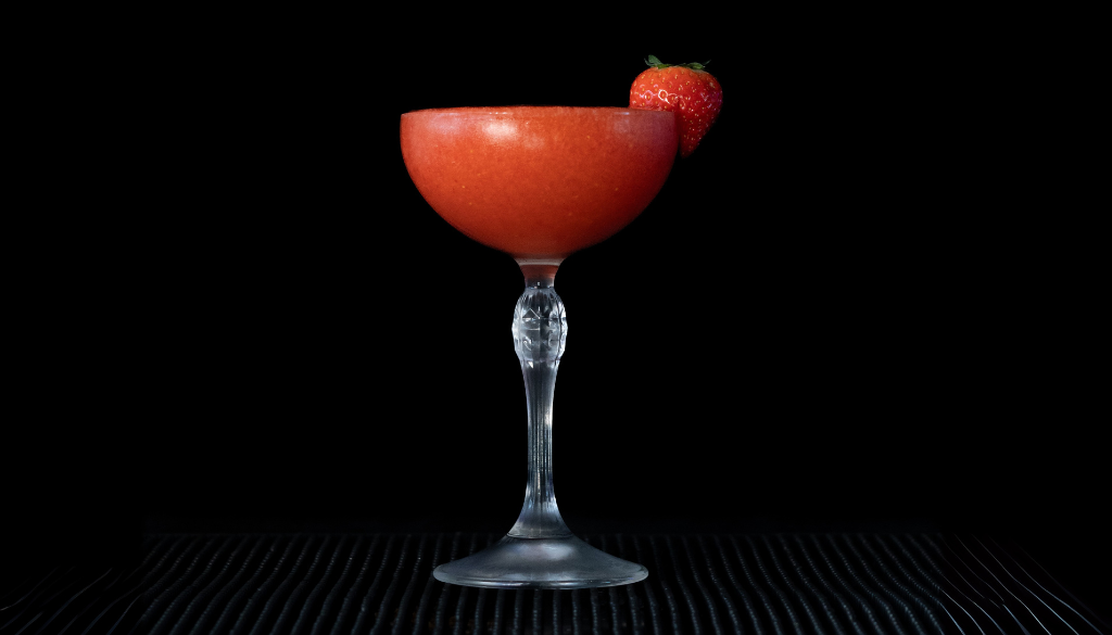 A Frozen Strawberry Daiquiri with a black background on a home bartop. The drink is a bright red and garnished with a fresh strawberry.