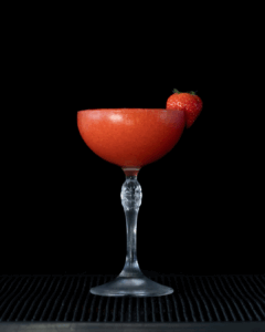 A Frozen Strawberry Daiquiri with a black background on a home bartop. The drink is a bright red and garnished with a fresh strawberry.