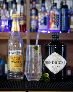 Hendricks & Fevertree Gin & Tonic with cucumber and next to a Hendricks Gin Bottle and a bottle of Fever-tree Indian Tonic Water on a home bartop.