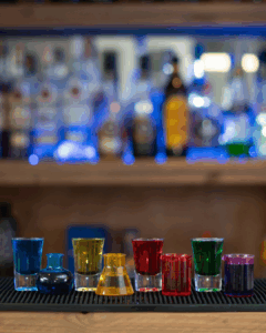 The Boo-zy Lab Kit, Halloween shots. Filled shot glasses with colored liquids. From left to right: Blue, yellow, red and green. Next to each shot glass is a mini test tube or Erlenmeyer filled with the same coloured shot mix. They are all sitting on top of a home bartop.