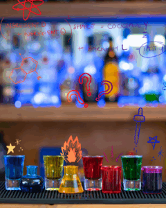 The Boo-zy Lab Kit, Halloween shots. Filled shot glasses with colored liquids. From left to right: Blue, yellow, red and green. Next to each shot glass is a mini test tube or Erlenmeyer filled with the same coloured shot mix. All the shots are on a home bartop.