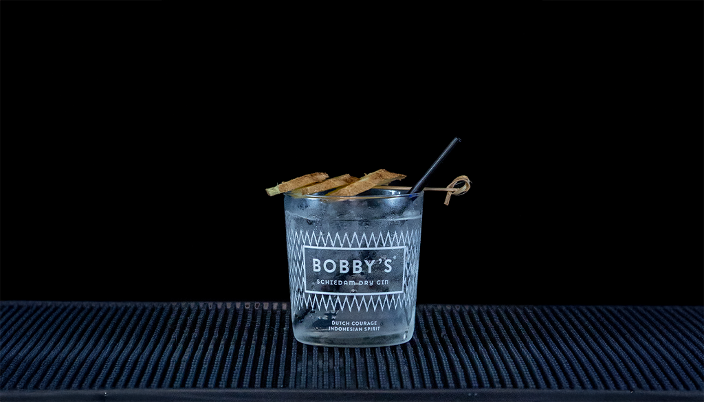 Bobby’s Pinang Raci Gin & Fever-Tree Indian Tonic in a glass with ginger and pepper on a home bartop with a black background.