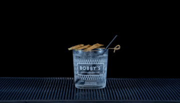 Bobby’s Pinang Raci Gin & Fever-Tree Indian Tonic in a glass with ginger and pepper on a home bartop with a black background.