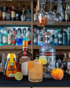 The Hot Honey Peach Margarita cocktail on a home bar top surrounded by its ingredients